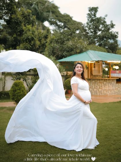 A dramatic outdoor shot featuring a mother-to-be in a white gown with a long, flowing train. The fabric catches the wind, creating a sense of freedom and beauty against the green backdrop.