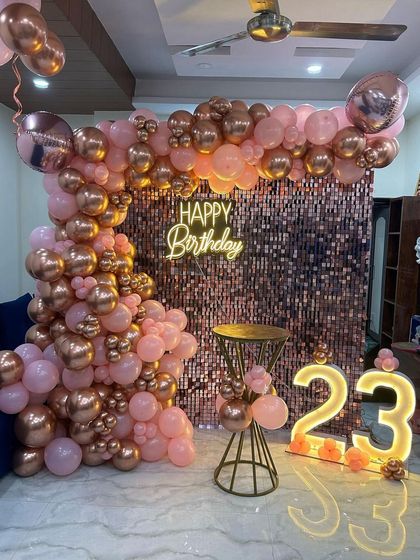 A lovely 23rd birthday setup in a home, using a rose gold sequin wall and a matching balloon arch. The marquee numbers and neon sign add a personalized and festive touch.