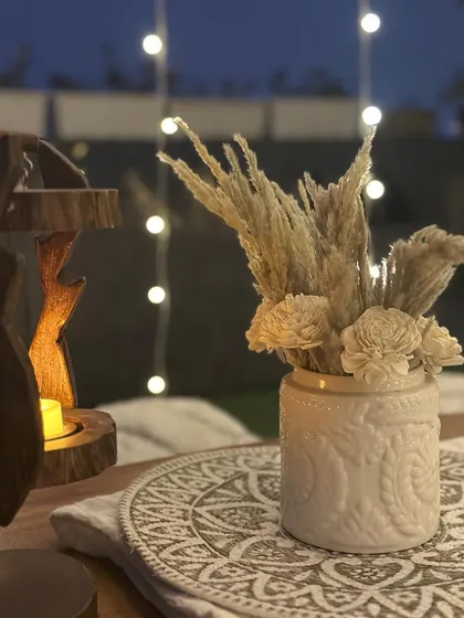 Cozy and boho vibes for a birthday soiree. This close-up shows the details of the tablescape, including dried pampas grass in a textured vase and a wooden incense holder, all against a backdrop of fairy lights.