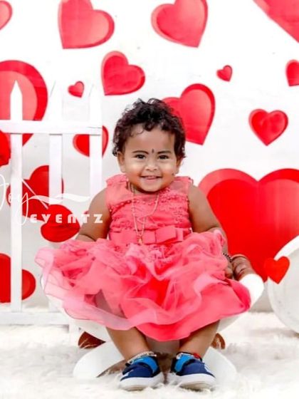 Celebrate love with a sweet Valentine's Day themed photoshoot. A baby girl in a lovely pink dress sits in front of a backdrop covered in red hearts, creating a cheerful and adorable picture.