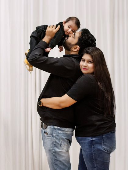 A joyful and candid family portrait. The father lifts the baby high in the air, creating a moment of pure happiness and playfulness, while the mother hugs him from behind.