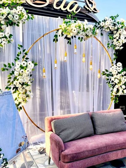 A beautiful outdoor Roka or engagement setup on a terrace. The golden ring frame with white florals, hanging bulbs, and a pink sofa creates a romantic setting against the open sky.