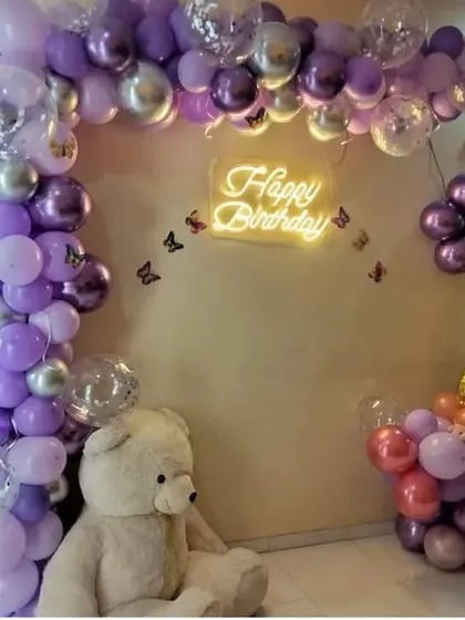 A cozy birthday setup at home with a purple and silver balloon arch, a teddy bear prop, and a 'Happy Birthday' neon sign.