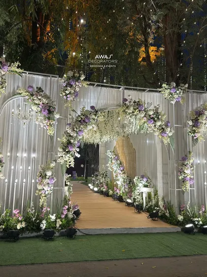 A dreamy entrance for an evening event, with white draped structures, arches of white and lavender flowers, and a ceiling of hanging wisteria.