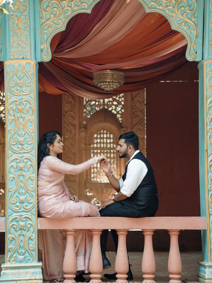 A romantic, regal-looking shot of a couple seated on a balcony of a palace-like set, dressed in beautiful traditional clothing.