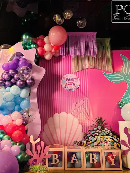 Dive into the fun with a detailed mermaid theme birthday party. This photo spot features a textured pink backdrop, a large mermaid tail cutout, coral props, and 'BABY' blocks, all surrounded by an ocean of colorful balloons.
