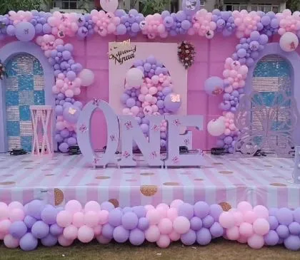For this first birthday, we went all out with a grand stage setup. The design includes a beautiful pink and lavender balloon arrangement framing custom-built backdrops, with large 'ONE' letters and the birthday girl's name as the centerpiece.