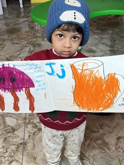 A toddler proudly displays his alphabet art, featuring a jellyfish and a jug of juice. We combine learning letters with creative drawing.
