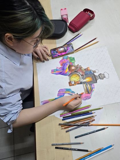 A student at her desk, surrounded by colored pencils, works on a vibrant still life composition, using a photo reference on her phone.