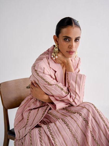 Styling for designer Esha Arora, this look features a pink striped pantsuit with delicate embroidery. The seated pose is elegant and thoughtful, highlighting the intricate details of the outfit.