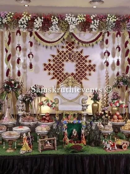 An exceptionally detailed and luxurious baby shower setup. The backdrop features an intricate geometric pattern made of flowers, complemented by a full display of offerings and traditional dolls.