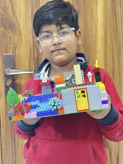 A student holds his LEGO model of a park scene with a small building and a blue water feature.