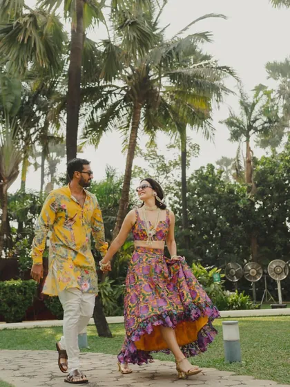 A candid shot of the couple walking hand-in-hand through a palm-fringed path during their Haldi event. Their vibrant, coordinated outfits and happy expressions set a joyful tone.