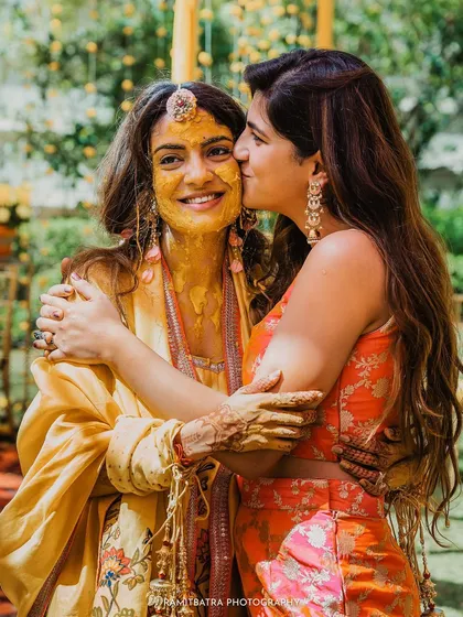 A sweet moment as the bride gets a kiss on the cheek from her sister or friend, her face glowing with Haldi.