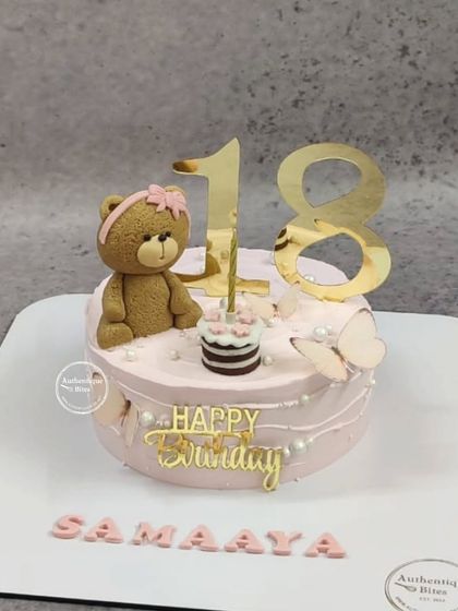 A sweet 18th birthday cake featuring a cute teddy bear. The pink cake is decorated with pearls, a butterfly, and a mini cake for the bear.