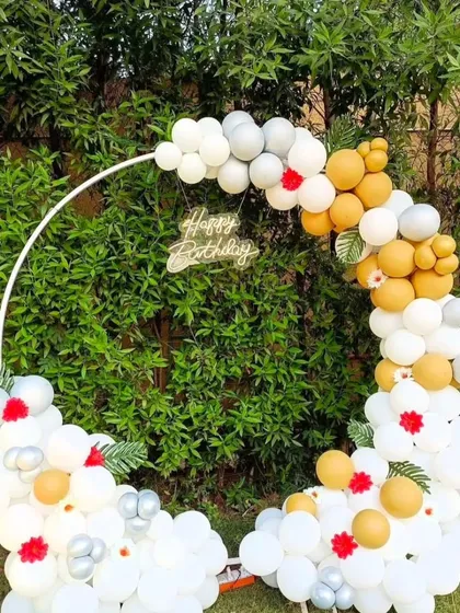 An elegant outdoor birthday backdrop. The circular frame is decorated with white, gold, and silver balloons against a natural green background.