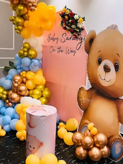 A cute teddy bear themed baby shower with a "Baby On The Way" message. This setup includes a large teddy bear cutout, a pink arched backdrop, and a colorful balloon arrangement in blue, yellow, and gold.