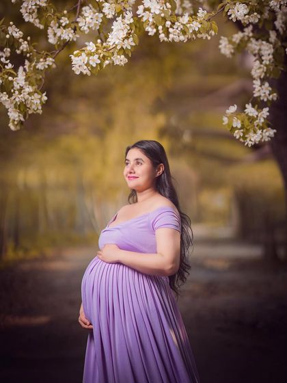 A serene portrait of a mom-to-be in a flowing lavender dress, standing under a canopy of white blossoms. The soft light and dreamy background create a peaceful and beautiful scene.
