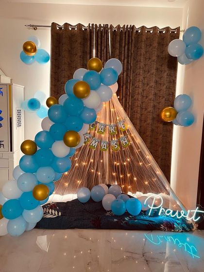A cozy canopy decoration in a living room. The tent is lit with fairy lights and framed by a blue, white, and gold balloon arch, with a custom neon name sign.