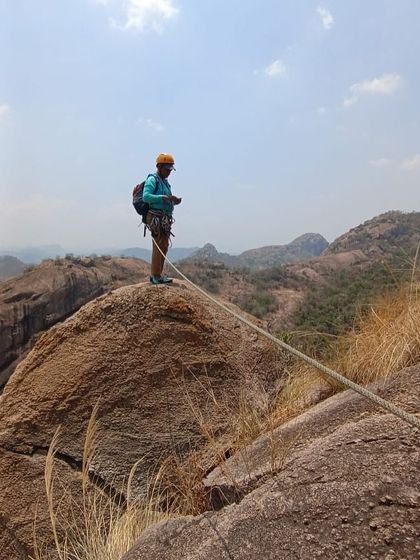A climber at a belay station high up on a multi pitch route, with a stunning view of the surrounding hills.
