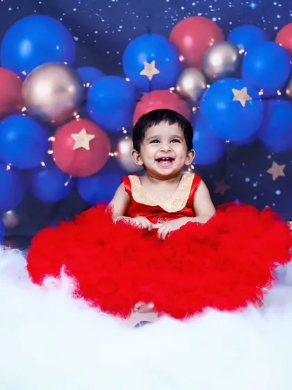 A magical fairy-themed shoot with a baby girl in a stunning red dress, sitting on a cloud under a canopy of balloons and stars.