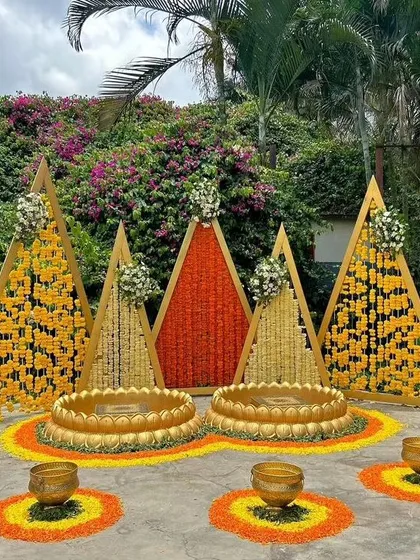 A stunning outdoor Haldi setup with triangular backdrops made of marigold and tuberose strings. The ground is decorated with floral rangoli, creating a picturesque setting for the ceremony.