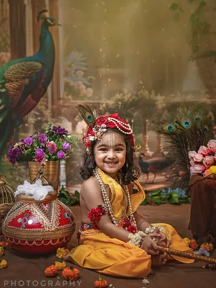 In my studio, I bring divine stories to life. This little one, dressed as Krishna with a playful smile, is surrounded by a rich, artistic setup featuring peacock feathers, marigolds, and a traditional makhan pot.