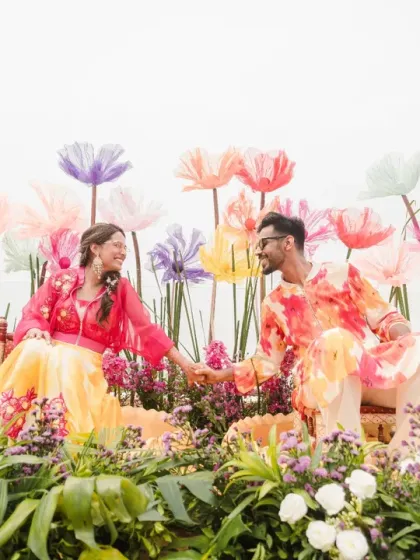 A vibrant and whimsical Haldi setup. The couple sits amidst a garden of oversized paper flowers, creating a surreal and beautiful scene.