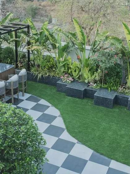 An overhead view of a terrace designed for both relaxation and entertainment. The layout includes a custom bar, a green lawn area, and checkerboard tiling, all surrounded by lush, tropical plants like the Traveller's Palm.
