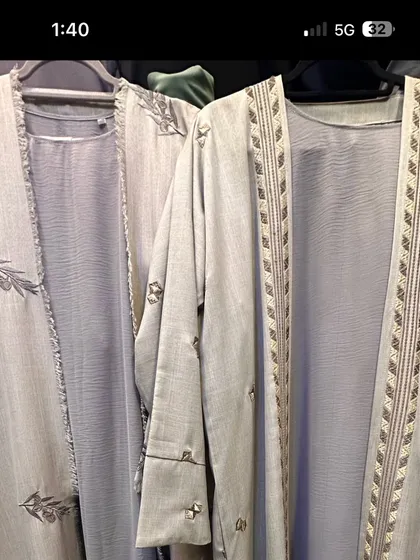 A pair of light grey open front abayas. One is detailed with a leaf pattern embroidery, while the other has a geometric design along the opening.
