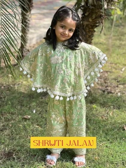A full look at the pistachio green cape set, complete with pom-pom trim and delicate embroidery. It's a fresh and unique look for any celebration.