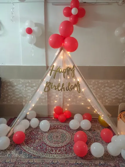 We offer cozy tent decorations for kids' birthdays at home. This simple and sweet setup includes a fabric tent illuminated with fairy lights, a 'Happy Birthday' banner, and red and white balloons.