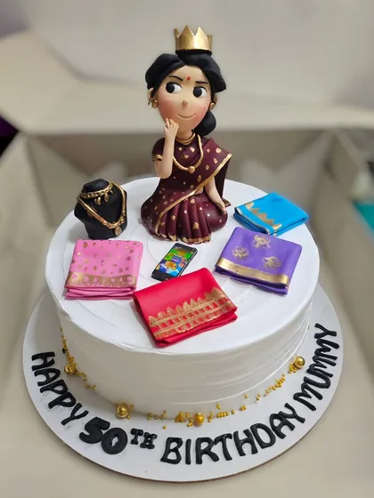 A beautiful cake for a 50th birthday, featuring a figurine of a lady who loves sarees, complete with miniature folded sarees and jewelry.