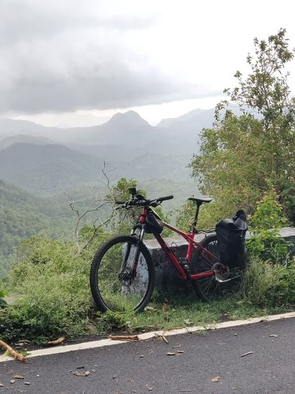 A mountain bike rests against a guardrail, overlooking the misty, layered peaks of the Palani Hills. This image shows the type of reliable rental bikes we provide, perfectly suited for the rugged terrain of our mountain tours.