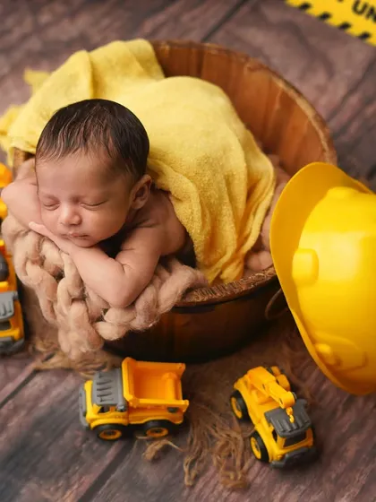 Caution: Cuteness under construction! This newborn is posed in a wooden bucket with a hard hat and toy trucks for a fun construction theme.