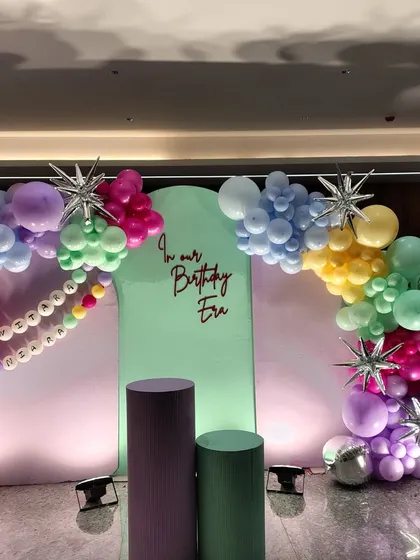 "In Our Birthday Era" - a fun and trendy pop star themed backdrop. The setup uses pastel arches and a vibrant rainbow balloon garland with star and disco ball accents, perfect for a music-loving birthday star.