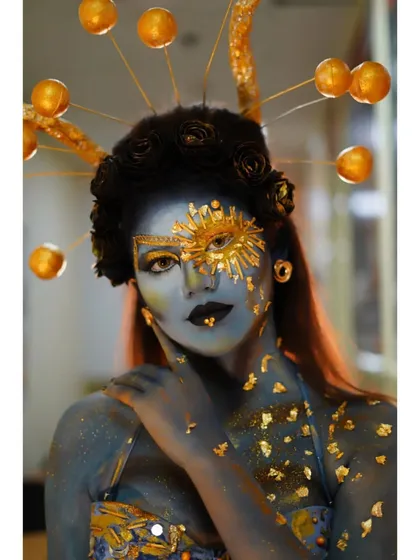 A celestial-themed creative look with blue body paint and golden accents, including a sun motif and a custom headpiece.