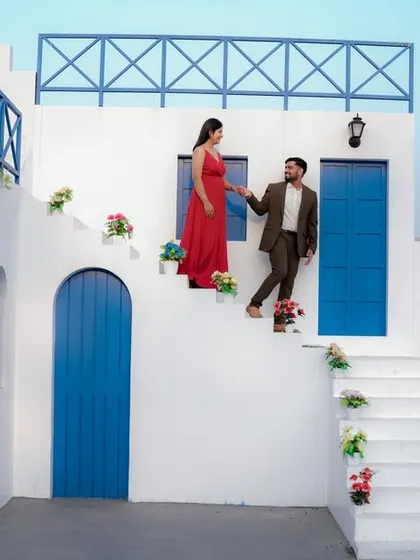 A fairytale moment on the steps of a beautiful Santorini-style building. The groom helps his bride-to-be, dressed in a flowing red gown, creating a classic and romantic cinematic shot.