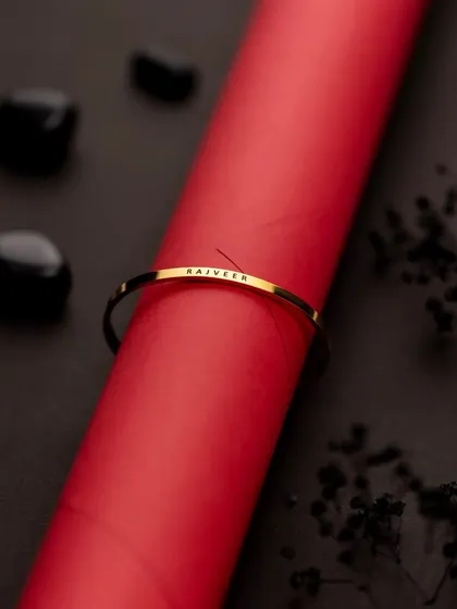 A creative product shot of a personalized gold bracelet against a bold red and black background. The composition is clean and graphic, making the product stand out.