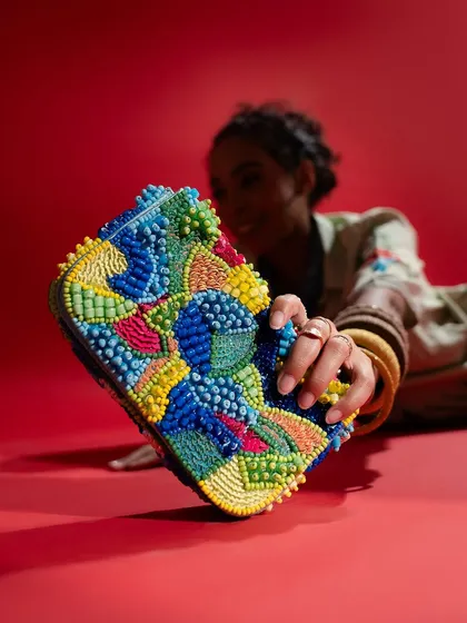 A close-up shot focusing on a unique, colorful beaded clutch. Even with the model slightly blurred in the background, the product remains the hero, showcasing its intricate detail and vibrant design against a bold red backdrop.