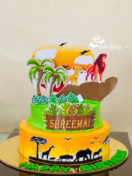 A vibrant two-tier Lion King theme cake. The design captures the spirit of the savanna with a sunset backdrop, jungle leaves, and silhouettes of the iconic characters.