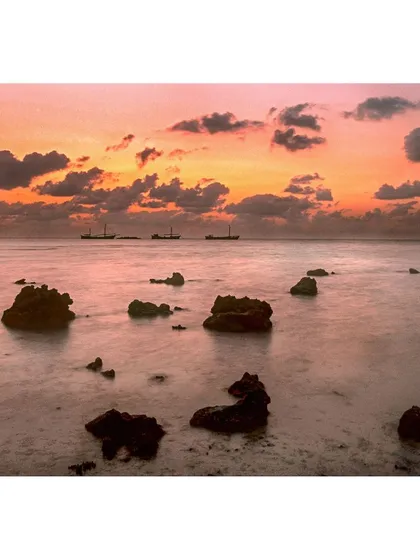 A tranquil seascape at dusk in Lakshadweep. The long exposure softens the water into a misty surface, contrasting with the sharp rocks and distant ships. This color landscape print captures the peaceful, painterly quality of the ocean at sunset.