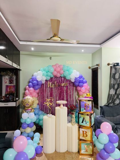 A lovely "Welcome Baby" decoration for a home, featuring a pastel balloon arch in mint, purple, and pink. The setup includes a shimmer backdrop, pedestals, 'BABY' blocks, and a cute teddy bear foil balloon.