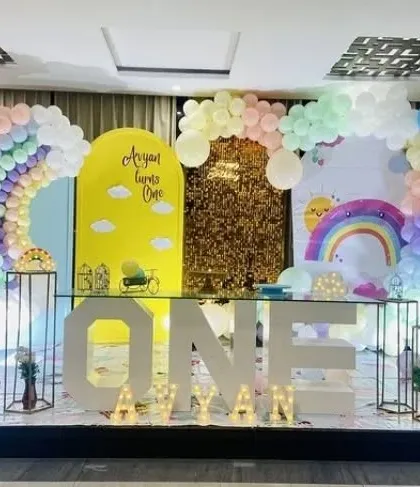 An elaborate and modern first birthday stage. This setup uses multiple backdrops, including a sequin wall and a rainbow arch, with large light-up 'ONE' letters and a glass table.