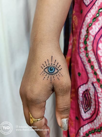 A small, colorful Evil Eye tattoo on the hand. This minimalist design serves as a modern talisman, offering protection with a pop of vibrant blue.