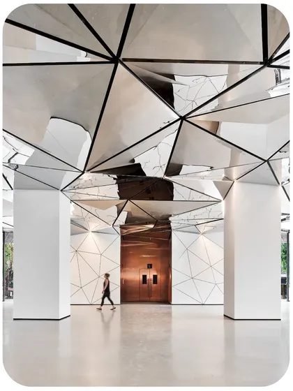 A view towards the rose-gold elevator lobby, where the triangulated ceiling continues, creating a seamless transition. The reflective surfaces capture and distort the surrounding geometry, making the space feel expansive and alive.