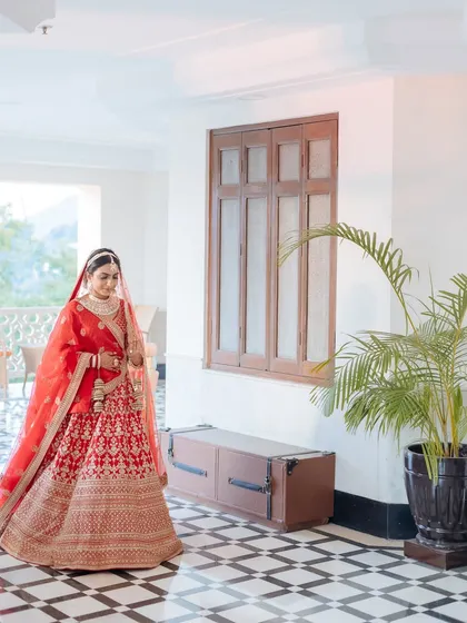A full-length portrait of a bride in her red lehenga, standing in a beautiful checkered-floor hallway.
