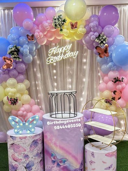 A pastel rainbow butterfly theme with a beautiful balloon arch and a "Happy Birthday" neon sign. The setup includes butterfly-themed plinths and decorative props for a magical photo opportunity.