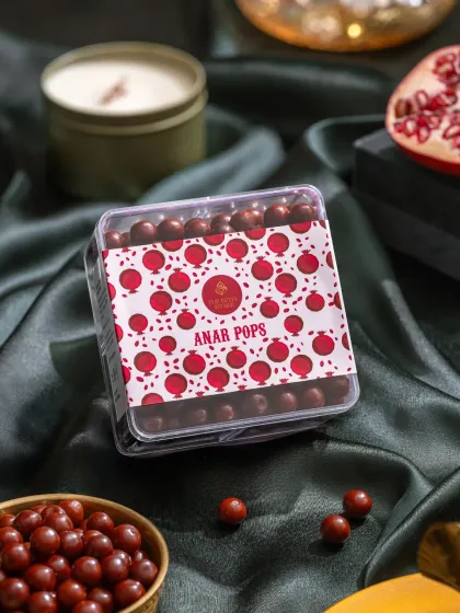 A box of Anar Pops (pomegranate candies), styled with fresh pomegranate and a candle for a warm, appealing look.
