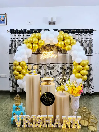 A simple and elegant home birthday decor in white and gold. The balloon arch, neon sign, and plinth arrangement create a clean and classic look.